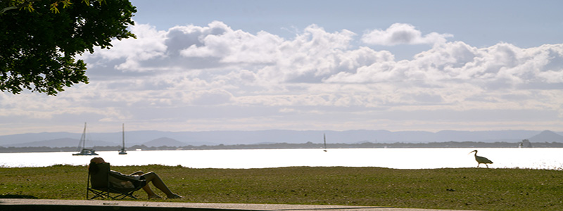 A person sitting on a camping chair facing Moreton Bay with boats and sea birds in the background.